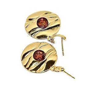 May include: A pair of gold earrings with a round, textured design and a small, red gemstone in the centre of each earring.