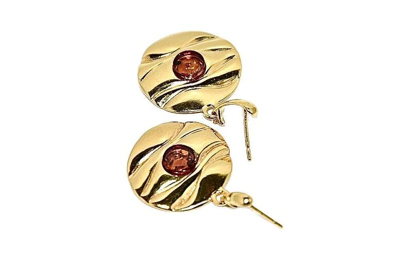 May include: A pair of gold earrings with a round, textured design and a small, red gemstone in the centre of each earring.