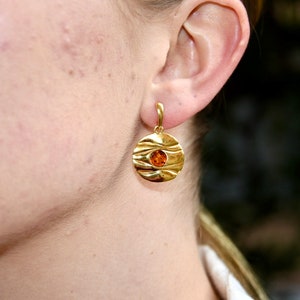 May include: Gold earrings with a round, textured design and a small amber stone in the centre.