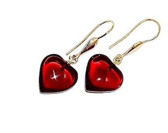 Puede incluir: Un par de pendientes en forma de corazón. Cada pendiente presenta un colgante de corazón rojo vibrante y translúcido, suspendido de un gancho dorado. Los pendientes están sobre un fondo blanco.
