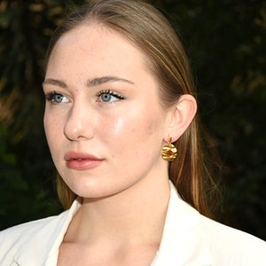 May include: A woman with long blonde hair wearing a white blazer and gold earrings with a red stone accent.