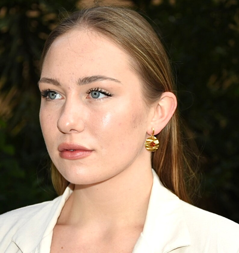 May include: A woman with long blonde hair wearing a white blazer and gold earrings with a red stone accent.