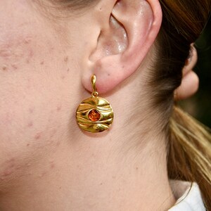 May include: Gold dangle earrings with a round, textured disc featuring a small orange gemstone in the centre.