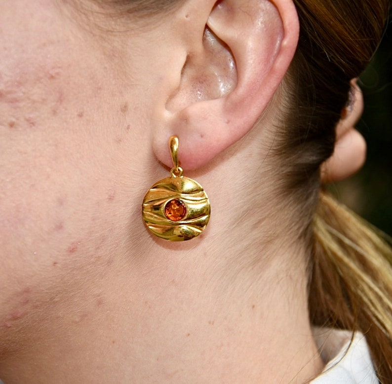 May include: Gold dangle earrings with a round, textured disc featuring a small orange gemstone in the centre.