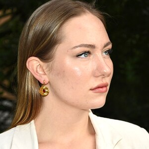 May include: A woman with long blonde hair wearing a white blazer and gold earrings with a red stone.