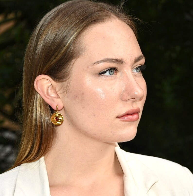 May include: A woman with long blonde hair wearing a white blazer and gold earrings with a red stone.