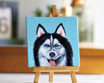 May include: A small square painting of a Siberian Husky dog on a miniature wooden easel. The dog has black and white fur, blue eyes, and a pink tongue. The background is a light blue color. The artist's name, @miniwindows, is printed on the easel.