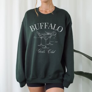 Può includere: Felpa verde scuro con la scritta bianca "BUFFALO Girls Club". Il design include un'illustrazione di due bicchieri da martini. La felpa è a girocollo.