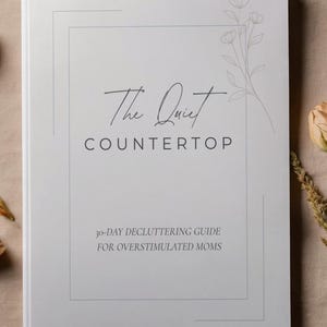 May include: A white book titled "The Quiet Countertop" with the subtitle "30-Day Decluttering Guide for Overstimulated Moms." The cover features a floral illustration and a minimalist design.