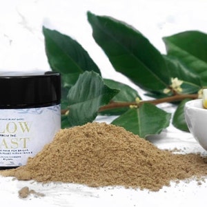 May include: A brown glass jar with a black lid containing a light brown powder. The jar has a white label with the text "Glow from the East Face Clay Mask for Bright Radiant Skin Filled with Vitamin C".