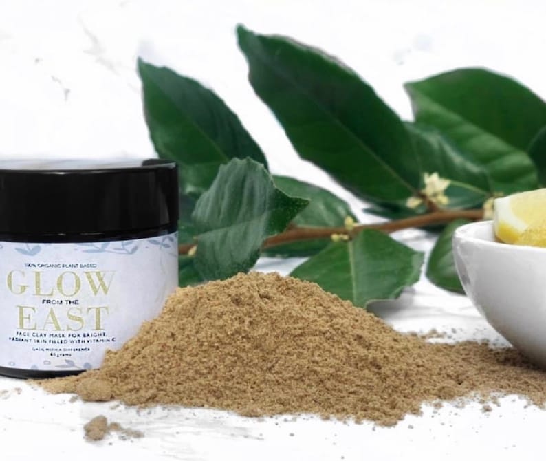 May include: A brown glass jar with a black lid containing a light brown powder. The jar has a white label with the text "Glow from the East Face Clay Mask for Bright Radiant Skin Filled with Vitamin C".