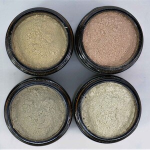 May include: Four small glass jars with black lids containing different shades of beige, light brown, gray, and light green powder.