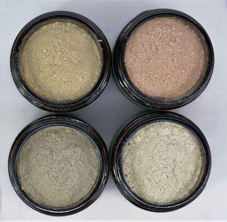 May include: Four small glass jars with black lids containing different shades of beige, light brown, gray, and light green powder.