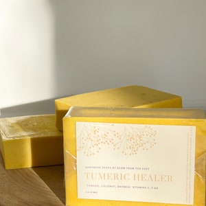 May include: Two bars of yellow soap wrapped in clear plastic. The top bar has a label that reads "HANDMADE SOAPS BY GLOW FROM THE EAST TUMERIC HEALER TUMERIC, COCONUT, OATMEAL VITAMINS C, E & B 3.5 OZ BAR".