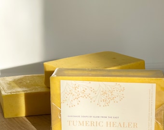 May include: Two bars of yellow soap wrapped in clear plastic. The top bar has a label that reads "HANDMADE SOAPS BY GLOW FROM THE EAST TUMERIC HEALER TUMERIC, COCONUT, OATMEAL VITAMINS C, E & B 3.5 OZ BAR".
