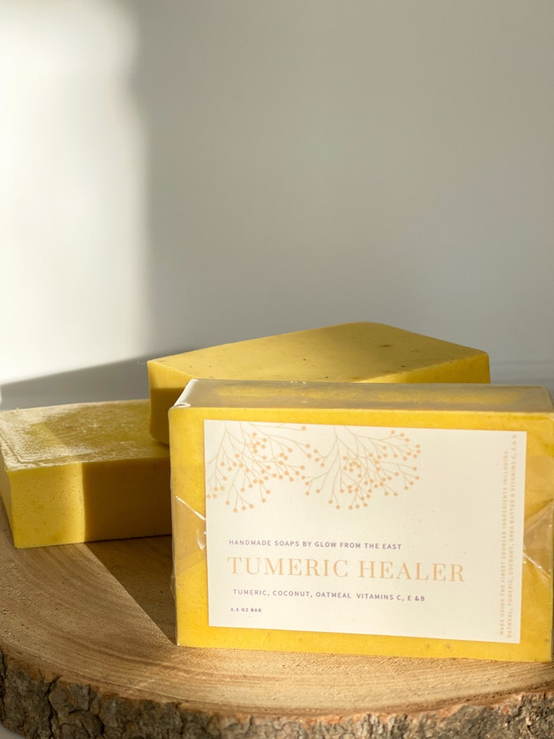 May include: Two bars of yellow soap wrapped in clear plastic. The top bar has a label that reads "HANDMADE SOAPS BY GLOW FROM THE EAST TUMERIC HEALER TUMERIC, COCONUT, OATMEAL VITAMINS C, E & B 3.5 OZ BAR".