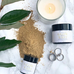 May include: Two jars of face mask powder labeled "Glow from the East" on a white surface with green leaves and two silver rings. The powder is a light brown color.