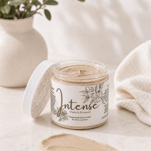 May include: A jar of "Intense" Chebe & Rosemary regenerating hair mask for Afro curly hair. The jar is open, revealing the mask's light brown color. A smear of the mask is next to the jar. A white lid, a white vase with greenery, and a white cloth are also in the image.
