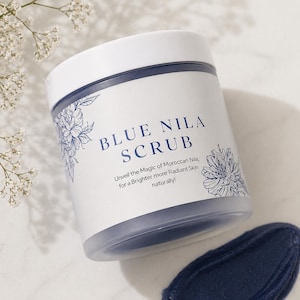 May include: A white jar of Blue Nila Scrub with a blue lid and floral design. The jar is next to a smear of the blue scrub. Text on the jar reads "Blue Nila Scrub" and "Unveil the Magic of Moroccan Nila, for a Brighter more Radiant Skin naturally!"