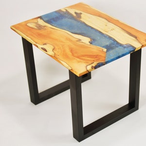 Pode incluir: Pequena mesa lateral de madeira com tampo retangular e pernas de metal preto. O tampo apresenta um padrão de grão de madeira natural com um rio de resina azul. A mesa mede aproximadamente 46 cm de comprimento, 30 cm de largura e 51 cm de altura.