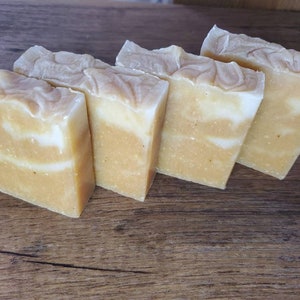 May include: Four rectangular bars of handmade soap. The soap has a layered design with a light yellow base and white swirls. The top of each bar has a textured, swirled pattern. The bars are arranged on a wooden surface.