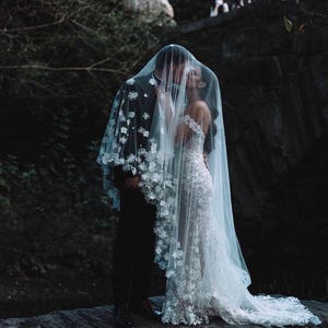 Op de afbeelding: Een stel omhelst elkaar onder een doorschijnende witte sluier versierd met bloemenapplicaties. De bruid draagt een witte kanten trouwjurk met een lange sleep. De bruidegom draagt een zwart pak. De scène speelt zich buiten af, met een donkere achtergrond.