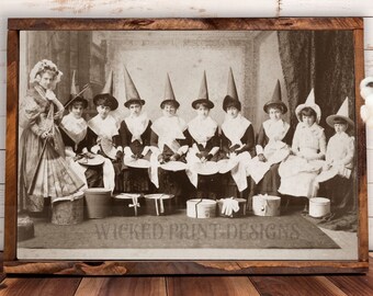 May include: A vintage black and white photograph of a group of women dressed in costumes, wearing witch hats and holding brooms. The women are sitting in a row, and there are several boxes in front of them. The photo is framed in a brown wooden frame.