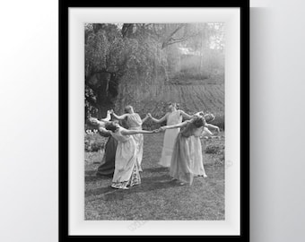 May include: Black and white photograph of five women in long dresses, holding hands and dancing in a circle in a grassy field.