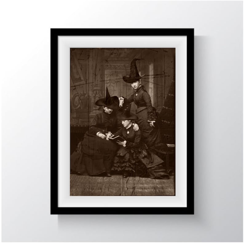 May include: A vintage black and white photograph of three women wearing black witch hats and long dresses. The women are sitting on a sofa and appear to be reading a book together.
