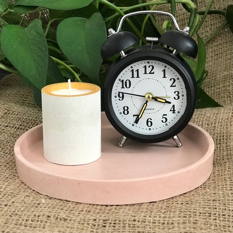 Pu&ograve; includere: Un vassoio in cemento rosa con una candela bianca e una sveglia nera con accenti argentati. Il quadrante dell'orologio ha numeri e lancette neri, e le parole "QUARTZ" stampate su di esso.