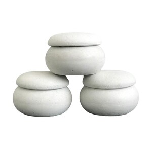 May include: Three white concrete lidded jars stacked on top of each other. The jars are round and have a smooth, slightly textured surface.