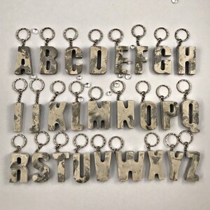May include: A set of 26 alphabet letter keychains made of concrete. Each letter is painted in a light grey colour with a marbled pattern. The keychains are attached to silver key rings.