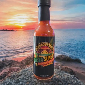 May include: A bottle of BASTA SUNSET Scotch Bonnet Hot Sauce, with a black cap and label. The label features the text "Underground Roots" and a tropical sunset scene. The bottle contains 5 fl oz (147 mL) of sauce.