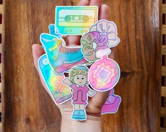 Puede incluir: Una mano sosteniendo una variedad de pegatinas holográficas de colores. Las pegatinas representan una cinta de casete, un patín, una niña de dibujos animados, una coleta, un teléfono y un sol con una cara sonriente.