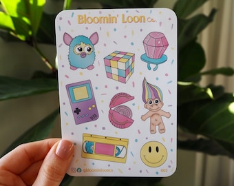May include: A sheet of stickers featuring a variety of 90s-themed designs, including a Furby, a Rubik's Cube, a troll doll, a cassette tape, a gameboy, and a smiley face. The stickers are on a white background with colorful confetti.