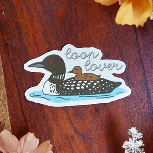 K&ouml;nnte beinhalten: Ein Aufkleber mit einem schwarz-wei&szlig;en Eistaucher mit einem Jungtier auf dem R&uuml;cken, das auf blauem Wasser schwimmt. Der Text "loon lover" ist in Kursivschrift &uuml;ber den Eistauchern geschrieben. Der Aufkleber hat einen wei&szlig;en Rand.