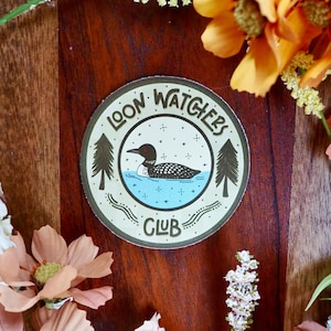 May include: A round sticker with the text "LOON WATCHERS CLUB" encircling a central image. The image features a loon swimming in blue water, flanked by two green pine trees. The sticker has a cream-colored background.