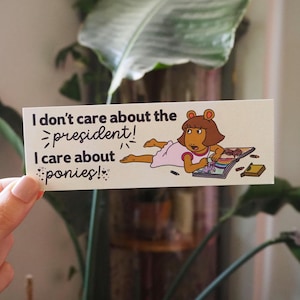 May include: A rectangular sticker with the text "I don't care about the president! I care about ponies!" and an illustration of a cartoon character in a pink dress. The character is lying down and drawing in a book.