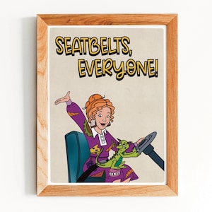 May include: A framed print with a cartoon illustration of a woman in a purple dress driving a bus, with a green dragon. The text "SEATBELTS, EVERYONE!" is displayed in yellow letters.