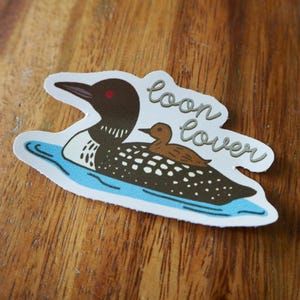 K&ouml;nnte beinhalten: Ein Aufkleber mit einer Cartoon-Illustration eines Eistauchers mit einem Baby-Eistaucher auf dem R&uuml;cken, der auf blauem Wasser schwimmt. Der Aufkleber tr&auml;gt den Schriftzug "loon lover" in Schreibschrift.