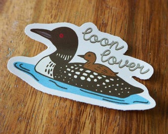 May include: A sticker featuring a cartoon illustration of a loon with a baby loon on its back, floating on blue water. The sticker has the words "loon lover" in cursive script.