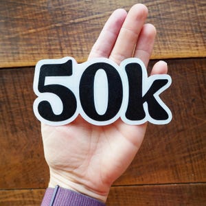 May include: A white sticker with the black text "50k" is held in a hand. The sticker has a black border and is on a wooden background. The person is wearing a purple long-sleeve shirt.