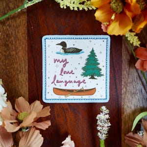 May include: Square sticker with a blue border and the words "my love language" in pink script. The sticker features a cartoon loon on water, a green pine tree, and an orange canoe with a paddle. The background is a dark wood surface with flowers.