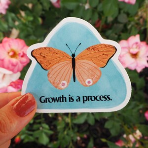 May include: A sticker featuring an orange butterfly with black details, set against a light blue background. The sticker has the text "Growth is a process." The sticker is held in front of a backdrop of pink roses.