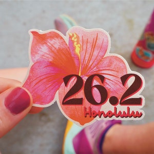 May include: A sticker featuring a vibrant hibiscus flower in shades of pink and orange. Bold, brown text reads "26.2 Honolulu," indicating a marathon distance. The sticker is held by a person, with a blurred background.