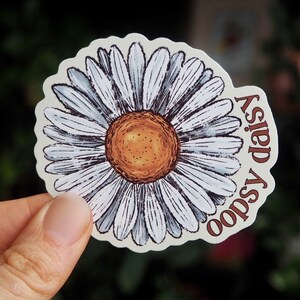 May include: A daisy sticker with a white and gray petal design and a brown center. The sticker has the words "oopsy daisy" in a brown, curved font. The sticker is held in a hand.