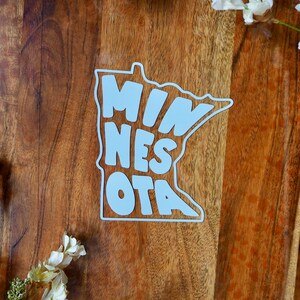 Könnte beinhalten: Ein weißer, konturgestanzter Aufkleber in Form des Bundesstaates Minnesota mit dem Wort MINNESOTA in einer fetten, serifenlosen Schrift. Der Aufkleber hat eine klare Umrandung und liegt auf einer dunklen Holzoberfläche.