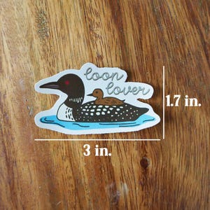 K&ouml;nnte beinhalten: Ein Aufkleber mit einer Cartoon-Illustration eines Eistauchers mit einem kleineren Eistaucher auf dem R&uuml;cken, der auf blauem Wasser schwimmt. Der Aufkleber ist 7,6 cm breit und 4,3 cm hoch, mit dem Schriftzug "loon lover".