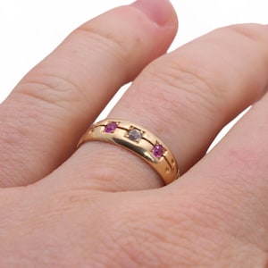 以下が含まれることがあります： 3つの宝石が付いたゴールドリング。リングの中央には透明な石があり、両側に2つの赤い石があります。バンドには装飾的なデザインがあります。リングは指にあります。