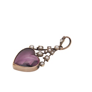 Puede incluir: Un colgante en forma de coraz&oacute;n con una piedra preciosa p&uacute;rpura facetada, enmarcada en oro. Peque&ntilde;as perlas redondas est&aacute;n dispuestas a lo largo de un tallo decorativo curvo. El colgante tiene un lazo para una cadena.